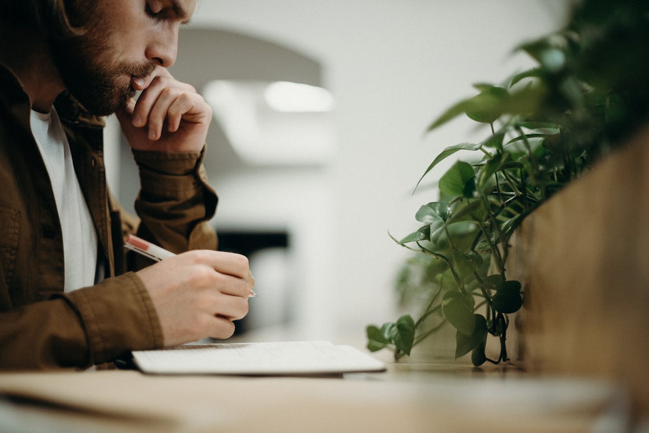 A man deeply concentrates while writing in an indoor office with plants.