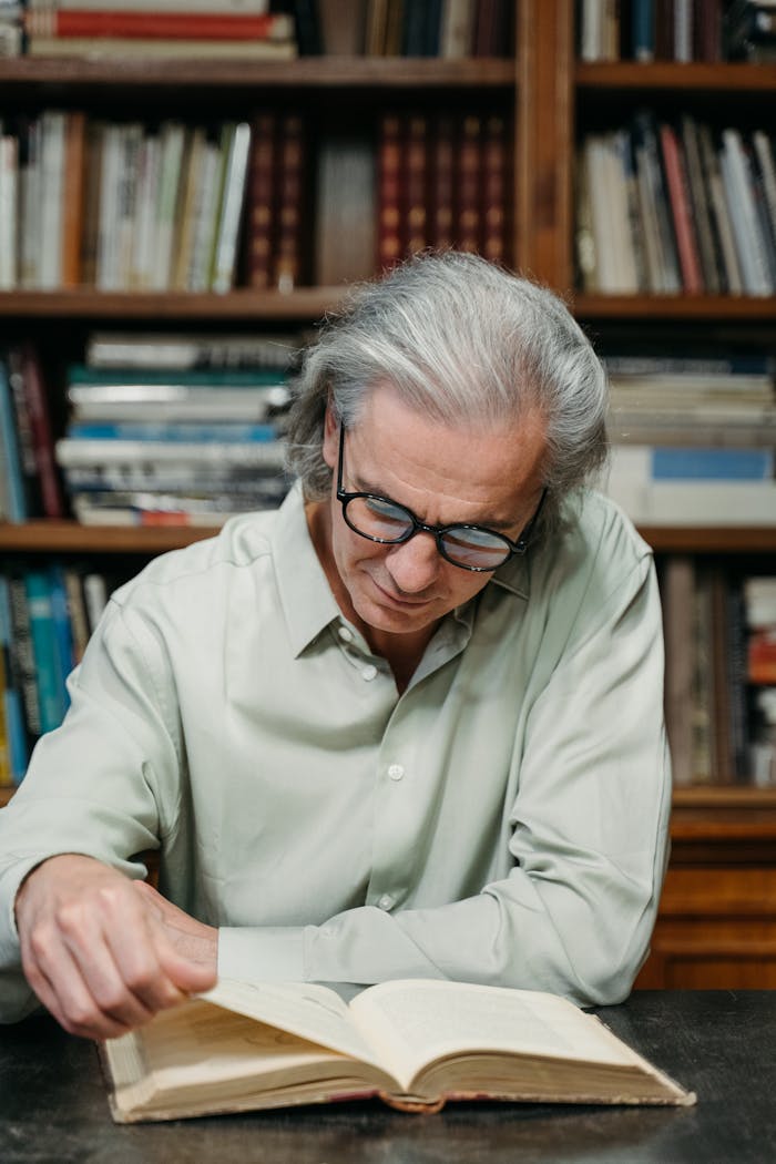Senior adult man with eyeglasses reading a book in a library surrounded by bookshelves.
